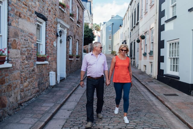 A couple dressed for summer walk towards camera down a narrow street.