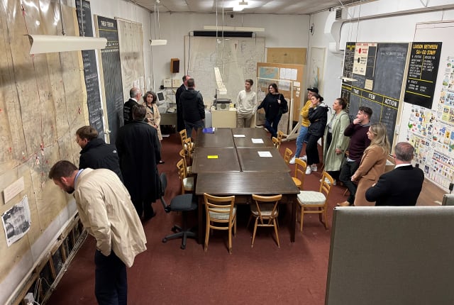 A large group of people stand around a long table with maps on the wall